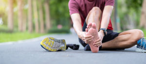 Male runner sitting on path with his sneaker off rubbing his painful heel