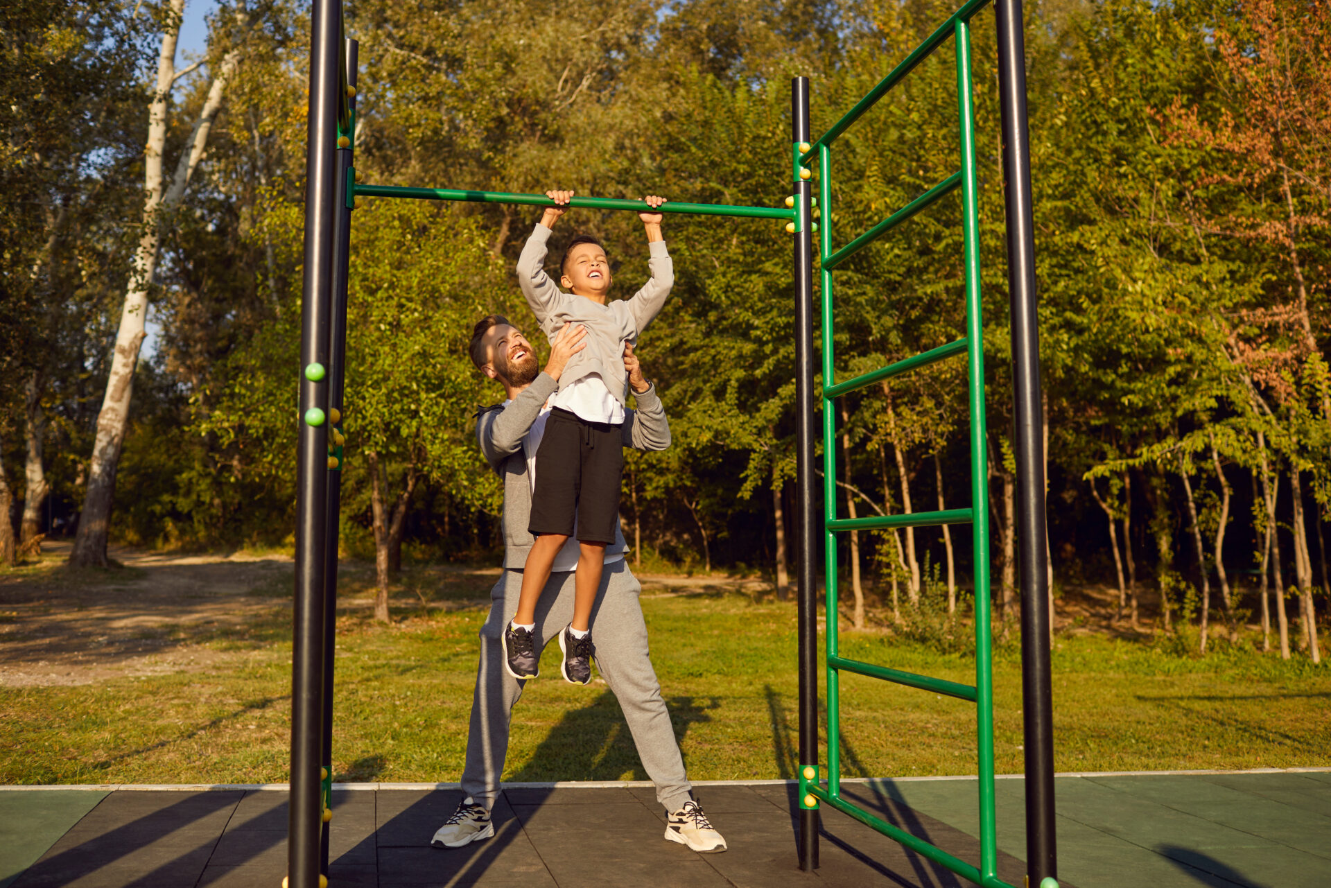 Young father or man coach teaching and helping a child boy to pull up himself on horizontal bar in summer park outdoors. Sporty family doing sport exercises in nature. Healthy lifestyle concept.