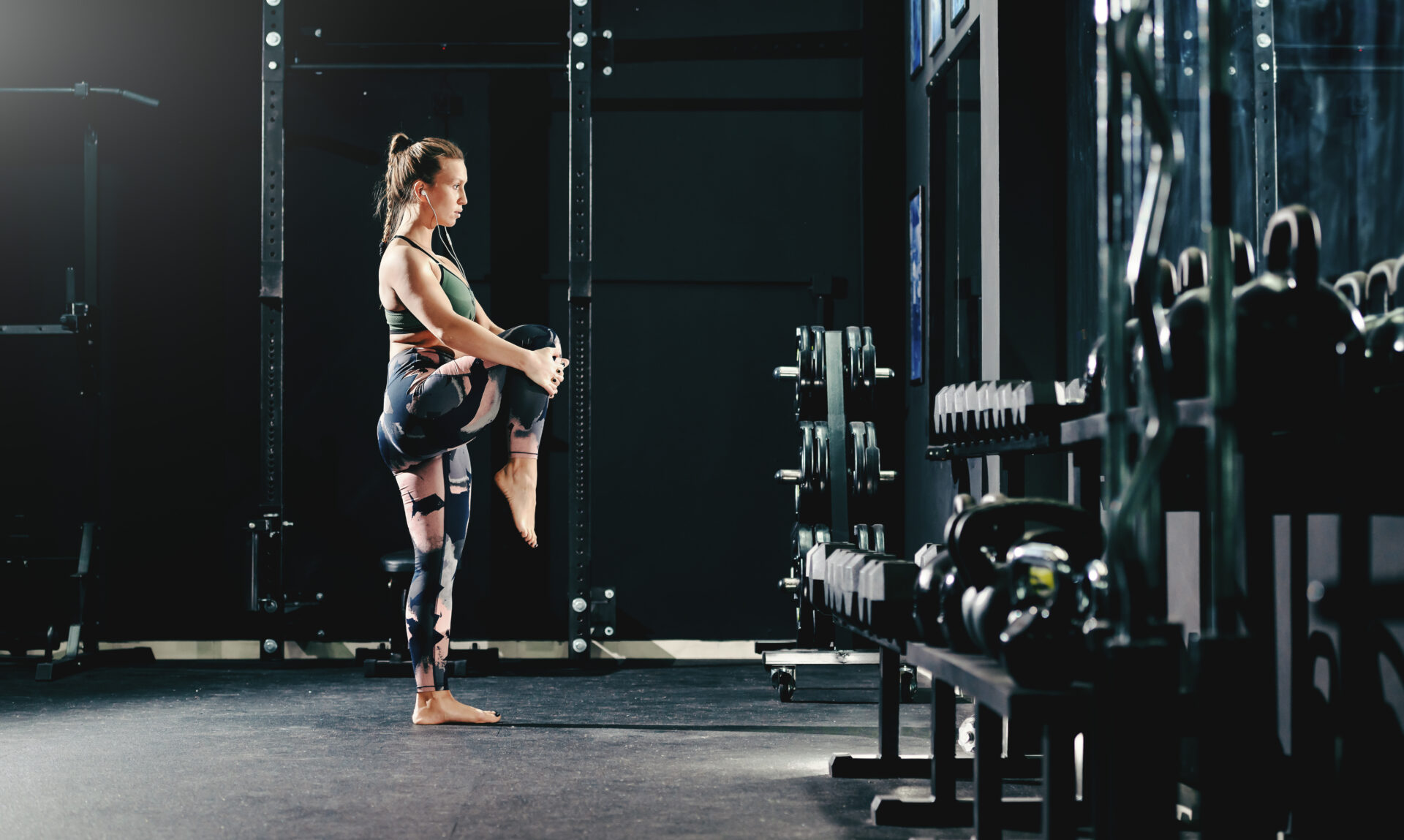 Side view of muscular woman in sportswear stretching leg while standing in gym at night. 