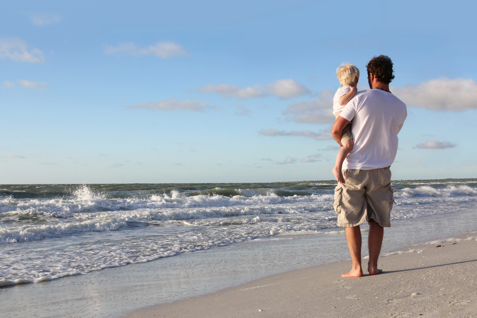 Father is holding his son in his arms during a walk on the white sand beach by the ocean. Father has recovered from a rotator cuff arthroscopy surgery.