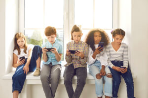 Group of 8-10 year old children sitting on windowsill, playing on their phones.